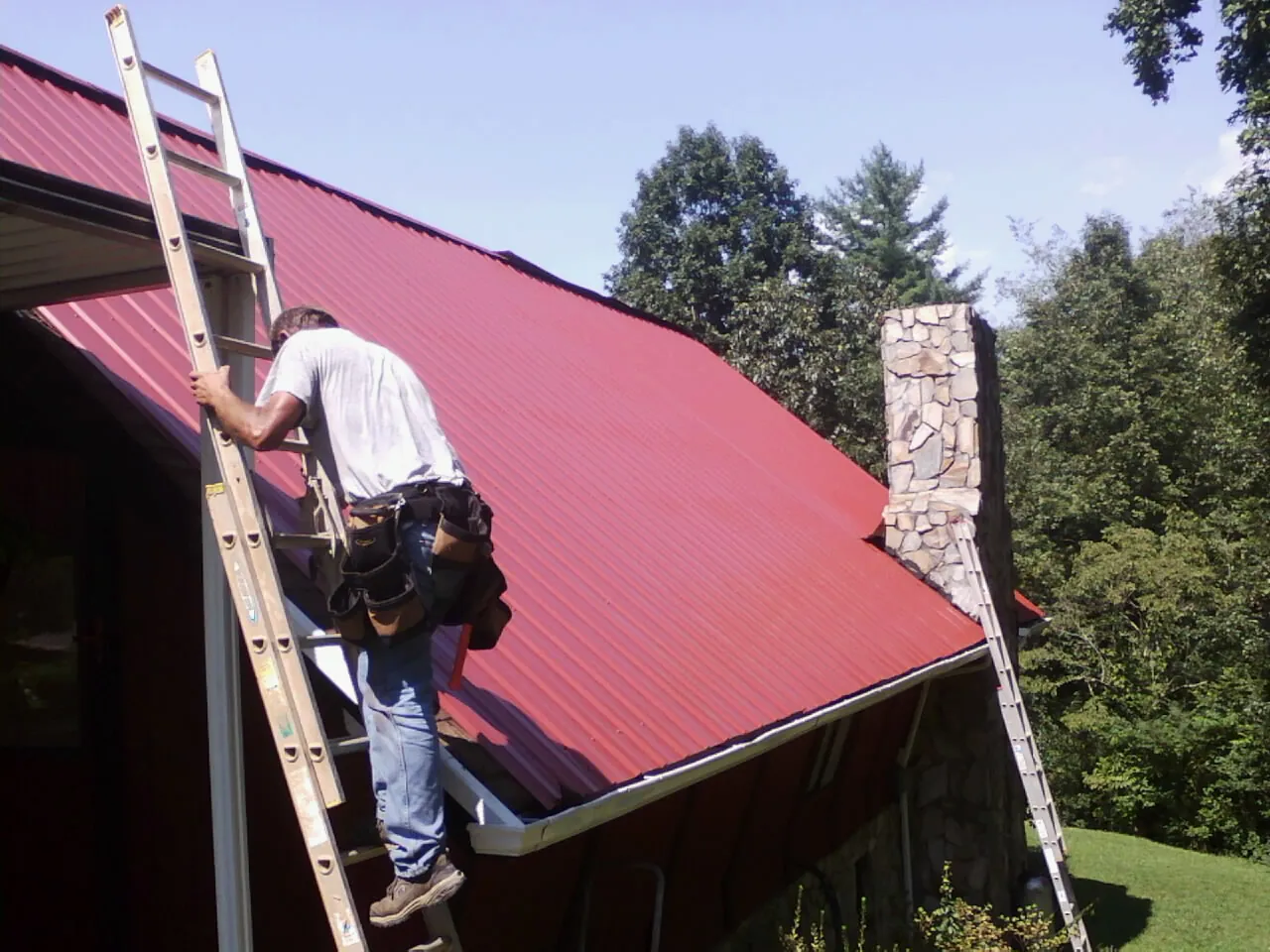 Roofer on a ladder beside a red metal roof and stone chimney — Soffit & Fascia Repair by Roofing Brothers in Gibsonia